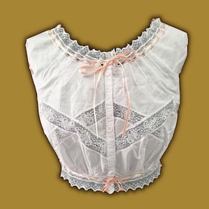 May include: White cotton blouse with lace trim and pink ribbon accents. The blouse features a scalloped neckline and a button-down front. The lace detailing is present on the bodice and hemline, creating a delicate and feminine aesthetic.