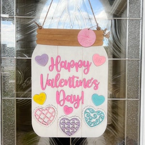 May include: A white wooden mason jar with the words "Happy Valentine's Day!" written in pink. The jar has a brown wooden lid and is decorated with colorful hearts and candy-shaped cutouts with phrases like "Sweet Talk" and "Too Cute".
