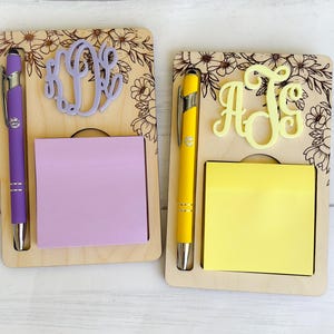 May include: Two wooden notepads with a pen holder and a personalized monogram. The notepad on the left has a purple pen and a lavender notepad. The notepad on the right has a yellow pen and a yellow notepad. Both notepads have a floral design.