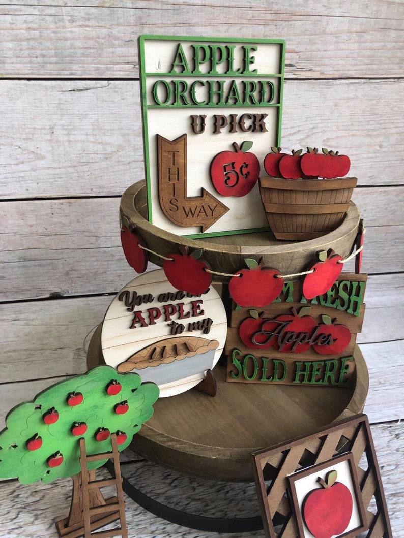 May include: A tiered wooden tray decorated with apple-themed wooden signs and decorations. The signs read "Apple Orchard U Pick", "You are the apple to my pie", "I am Fresh Apples Sold Here", and "This Way 5c". There is also a wooden apple tree with a ladder and a wooden basket of apples.