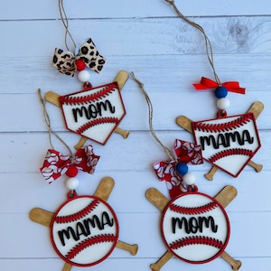 May include: Four wooden baseball ornaments with red and white baseballs and crossed bats. The baseballs have the words "Mom" and "Mama" written on them in black. The ornaments are decorated with red, white, and blue bows and beads.
