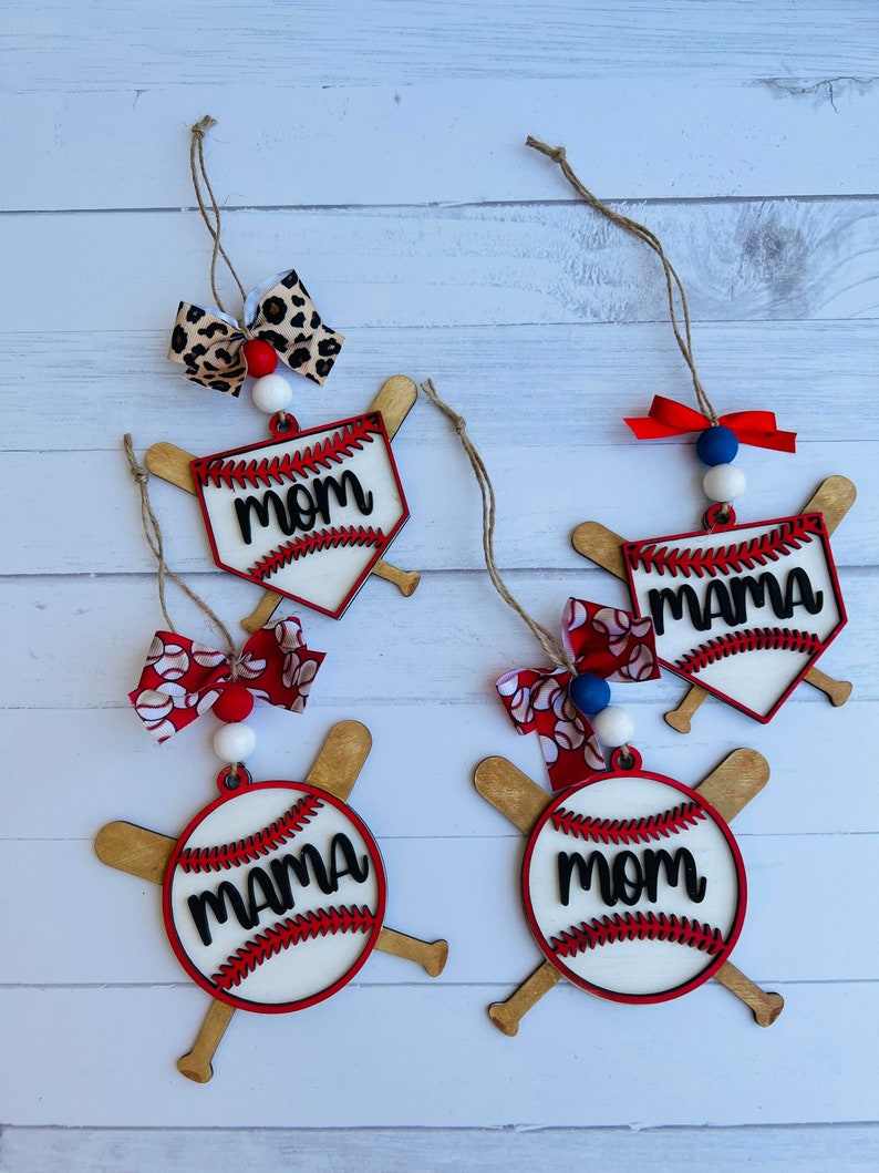 May include: Four wooden baseball ornaments with red and white baseballs and crossed bats. The baseballs have the words "Mom" and "Mama" written on them in black. The ornaments are decorated with red, white, and blue bows and beads.