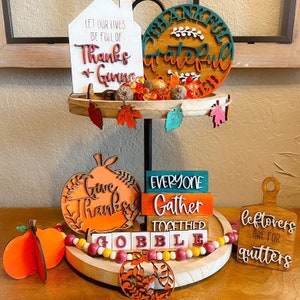 May include: A tiered tray decorated for Thanksgiving with wooden signs that say "Let our lives be full of Thanks + Giving", "Thankful Grateful", "Give Thanks", "Everyone Gather Together", "Gobble", and "Leftovers are for quitters". The tray is decorated with fall leaves, pumpkins, and beads.