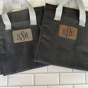 May include: Two dark gray tote bags with light gray handles. Each bag has a rectangular, brown leather patch with custom initials in a script font. One patch displays "SM" and the other displays "KDL".