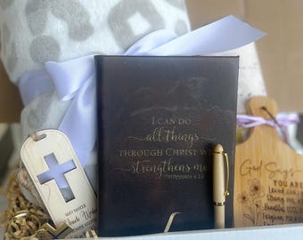 May include: A white gift box filled with a gray and white leopard print throw blanket, a black leather journal with the inscription "I can do all things through Christ who strengthens me Philippians 4:13", a wooden cross keychain, a wooden cross bookmark, and a wooden cutting board with the inscription "God Says You Are Loved John 3:16 Strong Phil 4:13 Beautiful Eccl 3:11 Forgiven Psalm 103:12".