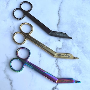 May include: Three pairs of candle wick trimmers in black, gold, and iridescent finishes. Each trimmer has a curved blade and double finger holes. The black trimmer is engraved with "Alexis", the gold with "Bailey", and the iridescent with "Mary".