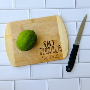 May include: A wooden cutting board with the text "SALT TEQUILA lime" engraved on it. A green lime is on the board, and a black handled knife is next to it.