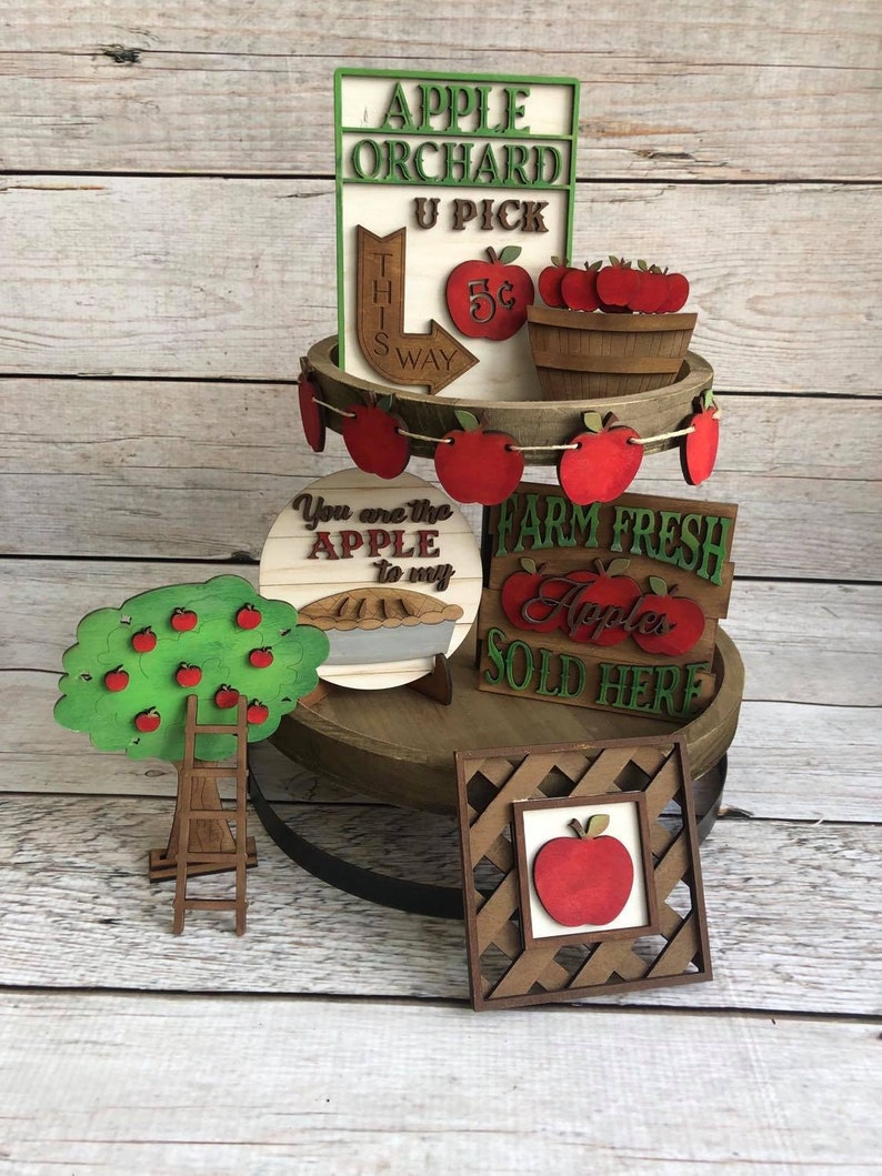 May include: A tiered wooden tray decorated with apple-themed wooden cutouts. The top tier features a sign that reads "Apple Orchard U Pick", a basket of apples, and a sign that reads "This Way 5&cent;". The middle tier features a sign that reads "You are the Apple to my", a pie, and a sign that reads "Farm Fresh Apples Sold Here". The bottom tier features a wooden apple tree with a ladder, and a framed apple cutout.