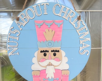 Puede incluir: Un letrero redondo de madera azul y blanco con el texto "Nuts About Christmas" en letras blancas. Un cascanueces rosa y azul con barba blanca está en el centro del letrero.