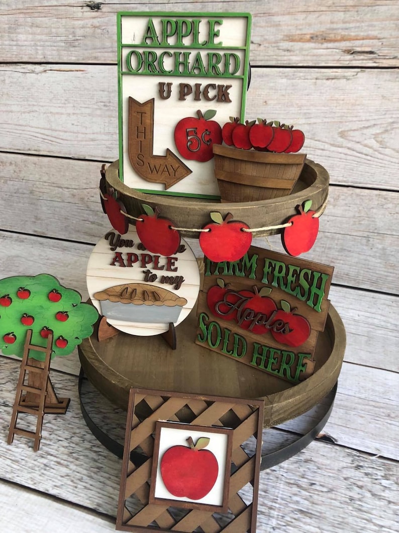 May include: A three-tiered wooden tiered tray decorated with apple-themed wooden signs. The top tier features a sign that reads "Apple Orchard U Pick" with a red apple and a sign that reads "This Way" with a red arrow. The middle tier features a sign that reads "You are an apple to my pie" with a pie and a sign that reads "Farm Fresh Apples Sold Here" with red apples. The bottom tier features a wooden frame with a red apple inside.
