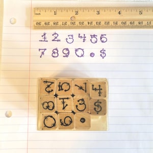 May include: A set of wooden number stamps in a clear plastic case. The stamps are arranged in a grid, with numbers and symbols printed in black ink. The image also shows handwritten numbers and a ruler with inch measurements.