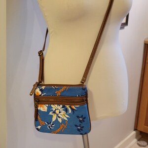 May include: A blue and white floral crossbody bag with a brown strap. The bag has two zippered compartments and the text "Love" and "Adventure" is printed on the front.