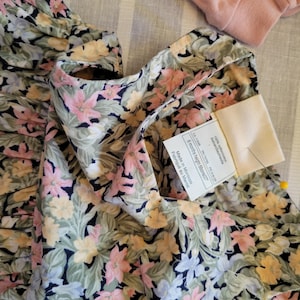 May include: A floral print garment with pink, yellow, and white flowers on a dark background. The garment has a ruffled edge and a tag that reads "6 months height 68cm/27". A pink garment is visible in the background.
