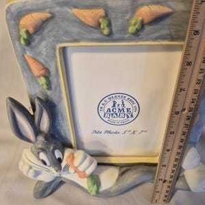 May include: A rectangular picture frame featuring a blue wash background with raised orange carrot accents and a Bugs Bunny figure at the base holding a carrot. The frame includes a Warner Bros. insert stating it fits a 5 inch by 7 inch photo.