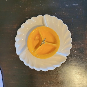 May include: A white ceramic serving dish with a yellow three-sectioned dish in the center. The dish has a metal handle in the center.