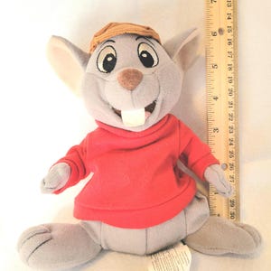 May include: A plush toy mouse with gray fur, wearing a red shirt and a brown cap. The mouse has large black eyes, a brown nose, and a big white tooth. A ruler is next to the toy, measuring up to 12 inches.