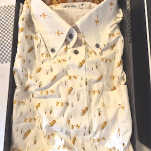 May include: A white button-down shirt with a whimsical pattern of skiers, trees, and arrows in shades of gold and green. The shirt is neatly folded in a black box. The collar and inner lining feature a brown cloud design.