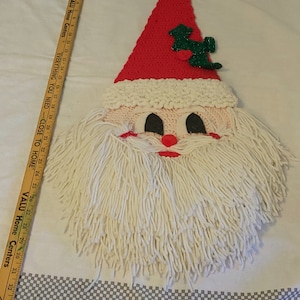 May include: A crocheted Santa Claus wall hanging with a red hat, white beard, and black eyes. The hat has a green holly sprig with red berries.
