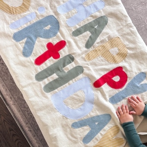 May include: A cream-colored fabric banner featuring the words HAPPY BIRTHDAY appliqued in various colors and patterns. The letters are crafted from blue, red, green, and yellow striped or solid fabric, stitched onto the base with visible thread.