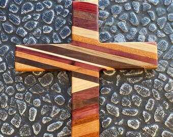 May include: A wooden cross made of various types of wood, with a striped pattern in shades of brown, red, and white. The cross is displayed on a gray textured surface.