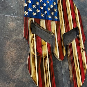 May include: Wooden Spartan helmet with an American flag design. The helmet is made of wood and is painted in the colours of the American flag. The helmet is a popular patriotic decor item.