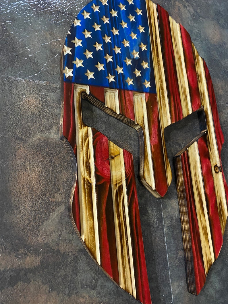 May include: Wooden Spartan helmet with an American flag design. The helmet is made of wood and is painted in the colours of the American flag. The helmet is a popular patriotic decor item.