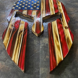 May include: A wooden Spartan helmet with the American flag painted on it. The helmet is made of multiple layers of wood and has a distressed look.