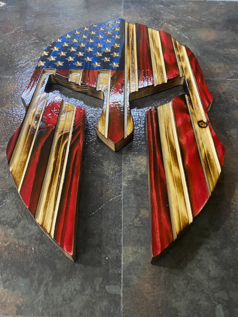 May include: A wooden Spartan helmet with the American flag painted on it. The helmet is made of multiple layers of wood and has a distressed look.