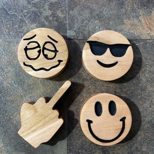May include: Four wooden emoji coasters featuring a smiley face, a smiley face with sunglasses, a sad face, and a middle finger emoji.