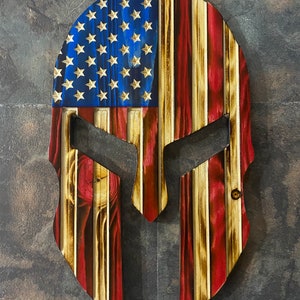 May include: A wooden Spartan helmet featuring an American flag design. The helmet is painted in red, white and blue with a wood grain texture.
