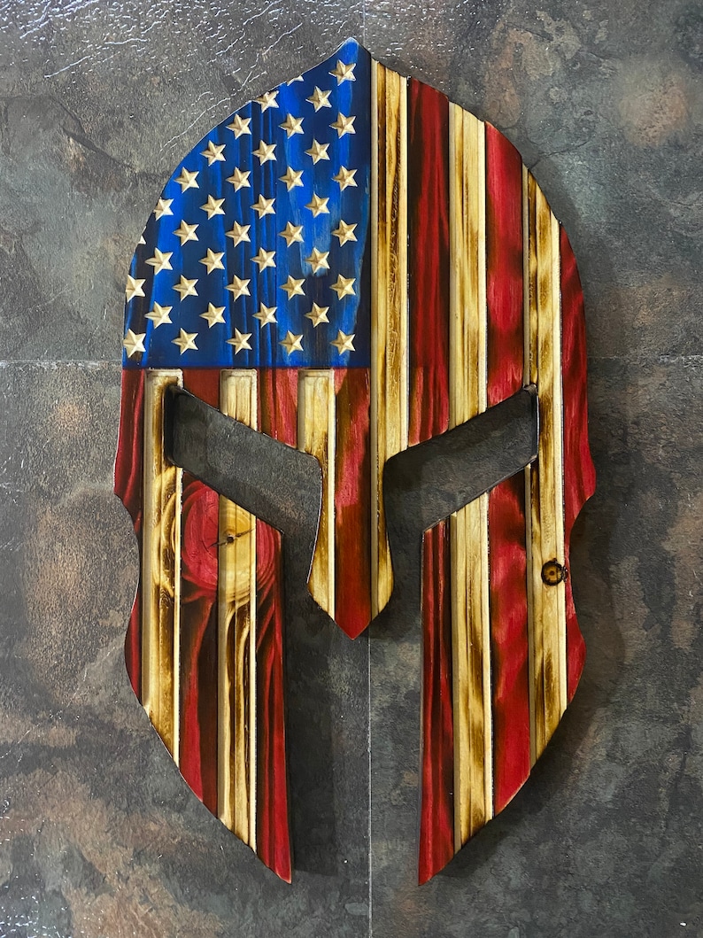 May include: A wooden Spartan helmet featuring an American flag design. The helmet is painted in red, white and blue with a wood grain texture.