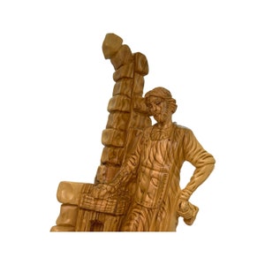 May include: A hand-carved wooden sculpture of a man carrying a large wooden beam on his shoulder. The sculpture is made of olive wood and has a natural, rustic finish.