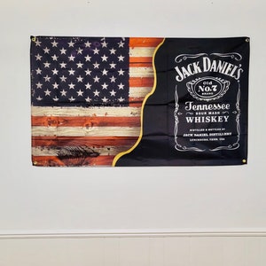 Può includere: Una bandiera nera e rossa con uno sfondo in legno a grana rozzamente lavorato. La bandiera presenta una bandiera americana sul lato sinistro e un logo Jack Daniel's sul lato destro. Il logo recita "Jack Daniel's Old No. 7 Brand Tennessee Sour Mash Whiskey Distilled & Bottled by Jack Daniel Distillery Lynchburg, Tenn. USA"