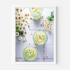 May include: Two glasses of green juice topped with white daisies on a white tiled surface.
