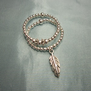 May include: Two silver beaded bracelets with a silver feather charm hanging from the bottom.