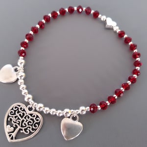 May include: A bracelet with deep red crystal beads and silver-coloured charms. It features heart-shaped pendants and a decorative tree of life charm. The bracelet's design is delicate and elegant.