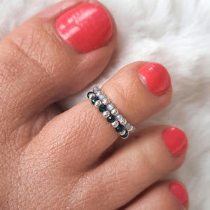May include: Two beaded toe rings. One ring features dark green beads, the other has clear beads. Both rings are silver in colour. The rings are worn on a toe with red nail polish.