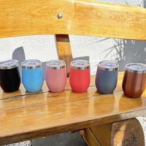 Könnte beinhalten: Ein Set aus sechs Edelstahlbechern mit transparenten Deckeln, die in einer Reihe angeordnet sind. Die Becher haben eine matte Oberfläche in Schwarz, Hellblau, Hellrosa, Korallenrot, Schiefergrau und Metallic-Kupfer und eine bauchige Form.