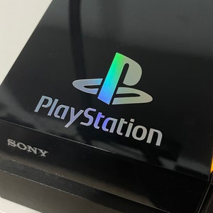 Könnte beinhalten: Eine schwarze PlayStation-Konsole mit einem holografischen PlayStation-Logo und dem Wort "SONY" in weiß.