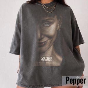 Può includere: Una t-shirt oversize grigio antracite con una stampa grafica del volto di una donna con un sorriso accennato. Il testo "JESSICA CHASTAIN OTHER MOMMY" e "YOUR MOMMY WON" è stampato sotto il volto. Il tessuto ha un aspetto vintage slavato.