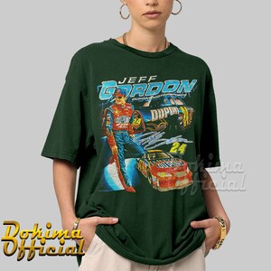 May include: A green t-shirt with a vintage-style graphic of Jeff Gordon, a NASCAR driver, in his race car. The graphic features the text "Jeff Gordon" and "Dupont Motorsports".