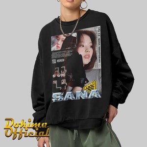 K&ouml;nnte beinhalten: Schwarzes Sweatshirt mit einem Grafikdruck eines Frauen Gesichts und dem Text "SANA MINATOZAKI" und "SANA". Die Grafik enth&auml;lt auch den Text "NO BAD VIBES" und ein gelbes Warnschild.