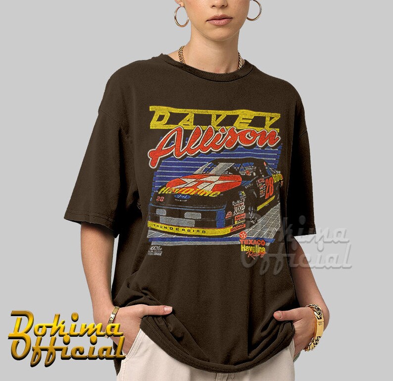 May include: Brown t-shirt with a vintage-style graphic of race car driver Davey Allison. The graphic features a red, white, and blue race car with the number 28 on the side. The text "Texaco Havoline Racing" is also visible on the graphic.