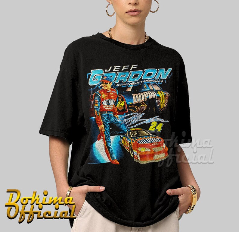 May include: Black t-shirt with a vintage-style graphic of race car driver Jeff Gordon, wearing a blue racing suit and a red helmet, standing next to his red race car with the number 24 on it. The text "JEFF GORDON" is printed in blue and white on the shirt.