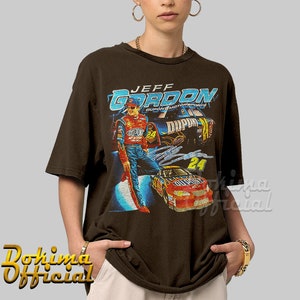 May include: A brown t-shirt with a vintage-style graphic of Jeff Gordon, a NASCAR driver, in his racing uniform. The graphic includes his name, the number 24, and the DuPont logo. The shirt has a crew neck and short sleeves.