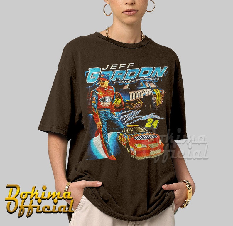 May include: A brown t-shirt with a vintage-style graphic of Jeff Gordon, a NASCAR driver, in his racing uniform. The graphic includes his name, the number 24, and the DuPont logo. The shirt has a crew neck and short sleeves.