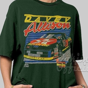 May include: A green t-shirt with a vintage-style graphic of race car driver Davey Allison. The graphic features a red, black, and white race car with the number 28 on the side. The text "Davey Allison" is in yellow and red lettering. The text "Texaco Havoline Racing" is also included in the graphic.