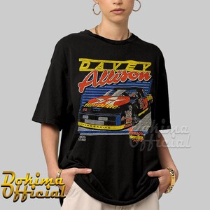 May include: Black t-shirt with a vintage-style graphic of race car driver Davey Allison. The graphic features a red, white, and blue race car with the number 28 on the side. The text "Davey Allison" is printed in yellow and blue above the car.