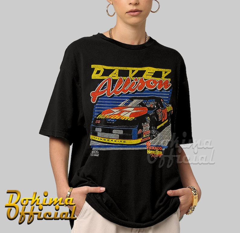 May include: Black t-shirt with a vintage-style graphic of race car driver Davey Allison. The graphic features a red, white, and blue race car with the number 28 on the side. The text "Davey Allison" is printed in yellow and blue above the car.