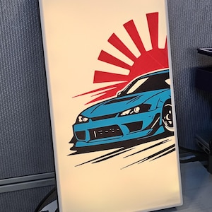 May include: A rectangular light panel featuring a graphic illustration of a blue sports car positioned in front of a red rising sun design. The artwork includes black motion lines beneath the vehicle, set against a cream-coloured background.